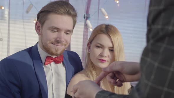 Elegant Young Blond Couple Sit in the Sofa and Hands of Other Male Is Counting Cards alt