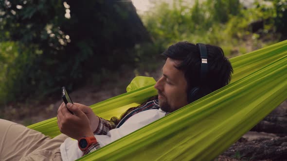 Man on Bicycle Trip at Camping By Lake is Relaxing in Green Hammock While Listening to Music alt