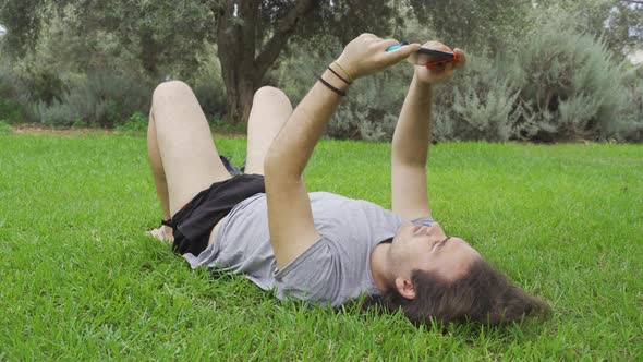 Man with long hair lying on the grass in the park and playing video games alt