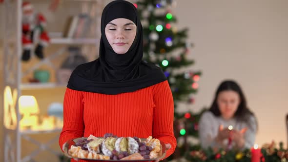 Young Happy Woman in Hijab Posing with Tasty Christmas Dessert in Living Room at Home As Blurred alt