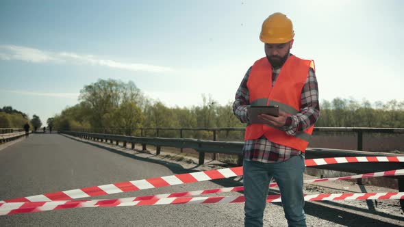Young Bearded Man Inspector in an Orange Vest and Protective Helmet with Tablet alt