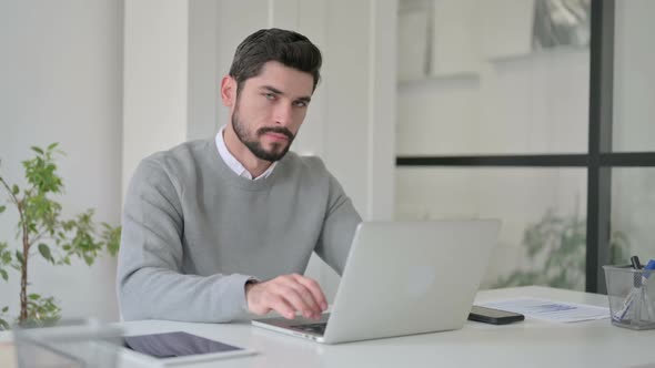 Young Man Pointing at Camera While Using Laptop in Office alt