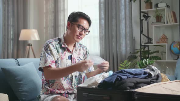 Asian Young Man Packing Clothes And Protect Face Mask In Suitcase At Home, Preparing For Vacation alt
