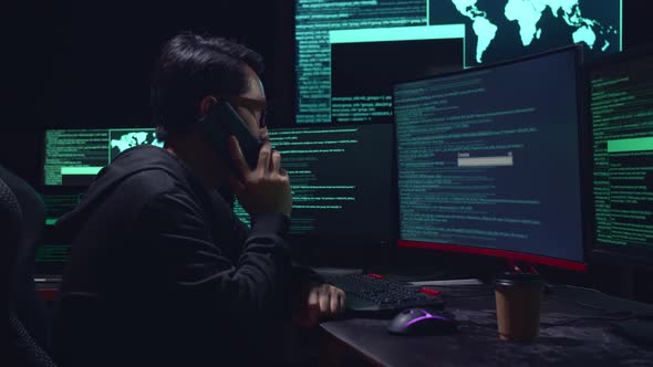 Asian Male Hacker Talking On Phone With Multiple Computer Screens And Success Downloading Data alt