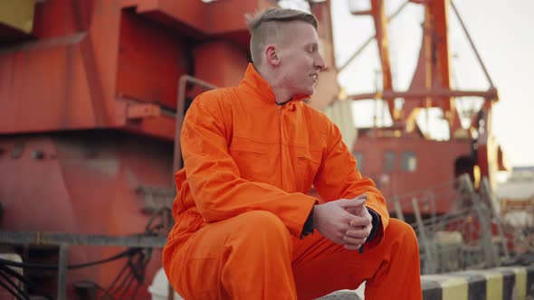 Young Man in Orange Uniform Sitting During His Break By the Sea in the Harbor alt
