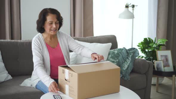 Beautiful Mature Woman Open Cardboard Box Sitting on Couch in Living Room. alt