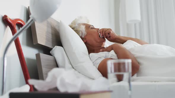 Sick african american senior woman holding tissue coughing while lying on the bed at home alt