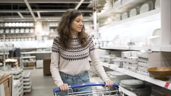 Young Woman Rolls Shopping Cart Along Shelves with Goods in Supermarket in Kitchen Department alt