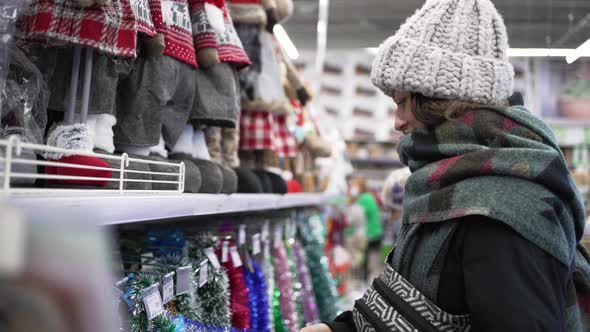 A Young Woman in a Black Down Jacket in a Supermarket Chooses Christmas Decorations alt
