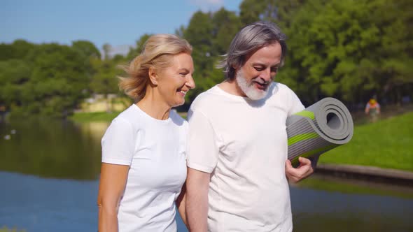 Lovely Senior Man and Old Woman Holding Yoga Mat for Exercising Outdoor Walking in Park alt