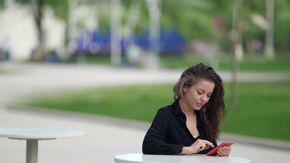 Woman with Smartphone in Street Cafe in Park Area at Summer Day Sending Messages in Social Nets alt