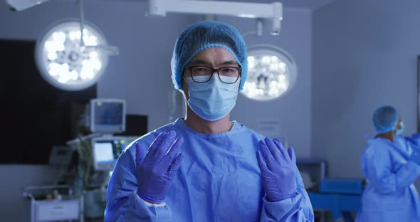 Portrait of asian male surgeon wearing face mask, gloves, cap and scrubs in operating theatre alt