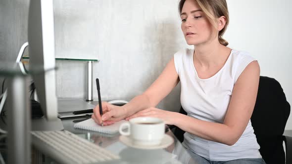 Young Woman Student Sitting in Front of Computer with Coffee Learns Online Writes Notes in Notebook