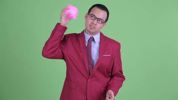 Stressed Asian Businessman with Eyeglasses Holding Piggy Bank and Giving Thumbs Down alt