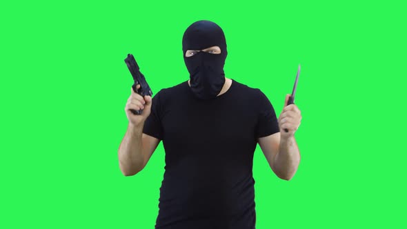 A man in a black mask holds a knife and a gun in his hand.Balaclava.Green screen background.