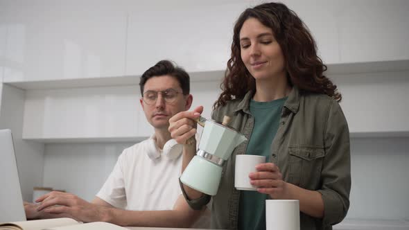Caring Wife Pours Coffee for Her Husband While He is Working on a Laptop Sitting in the Kitchen alt