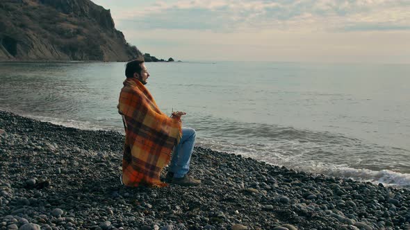 Slow Motion Young Man with a Beard and Mustache Sits in a Blanket and Coughs on a Deserted Seashore alt