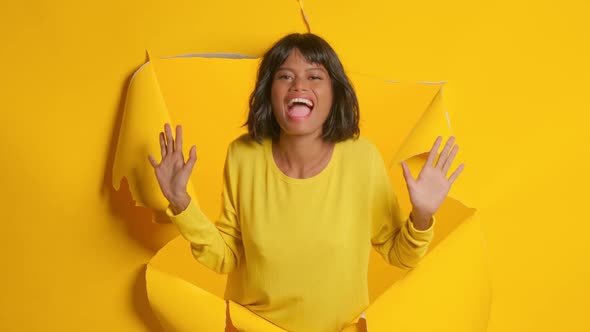 Young Woman Exclaim with Joy Raises Hands in Ripped Hole Yellow Background alt