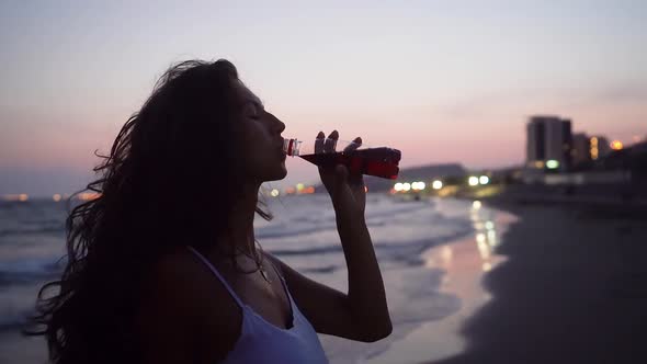 Woman Drinking Soda on Beach with City Lights in Background