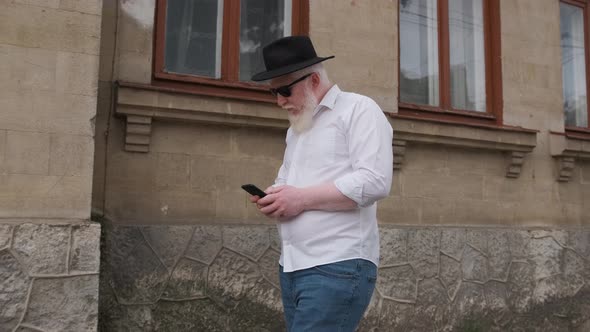 Old Man with Albinism Browsing on the Internet While Walking Next To Old House alt