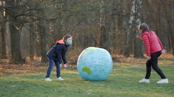 Two Girls in Medical Masks Play in the Spring Park with a Large Inflatable Ball Planet Earth. The