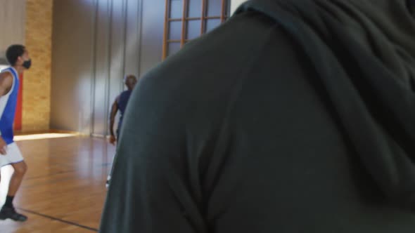 Diverse male basketball team playing match with coach holding clipboard, wearing face masks alt