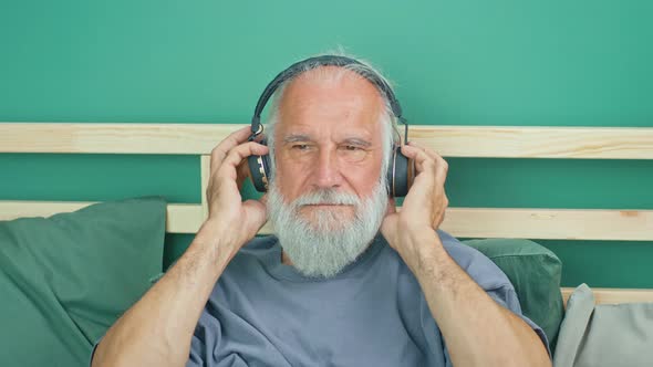 Carefree Elderly Man Listening to Music With Big Headphones on the Bed at Home alt