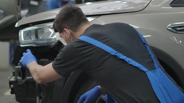 Back View of Auto Mechanic in Coronavirus Face Mask Sitting at Broken Vehicle in Repair Shop alt