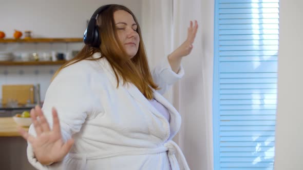 Cheerful Fat Woman Listening To Music in Headphones and Dancing Near Window at Home alt