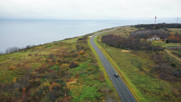 Drone Flying Above Black Car Moving Along Clear Road Near Beautiful Lighthouse Sea Bay and Autumn alt