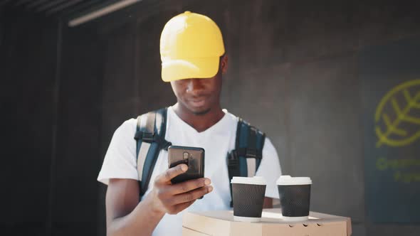 African Delivery Man with Package and Smartphone While Carrying a Box with Pizza alt