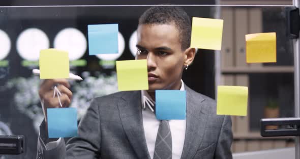 Young African Man in a Modern Office, Draws Arrows Infographic on the Glass Board with Sticky Notes alt