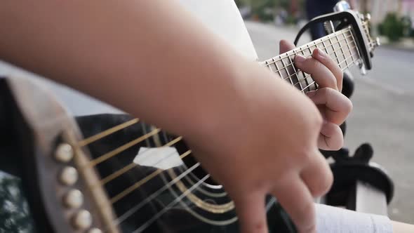 Hands of a Teenager Boy Holding an Acoustic Guitar Play Outdoors in the Evening on a Bench alt