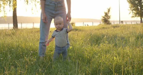 Infant Standing Uncertainly Holding Mother's Hand and Looking at Camera Outdoors alt