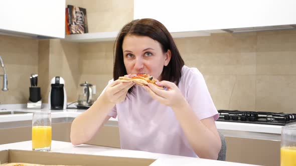 Young Woman Eats Slice of Italian Pizza Sitting in Kitchen