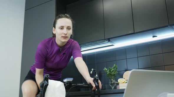 Young female drinking water from bottle while training and pedaling on stationary smart bicycle alt