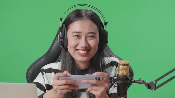 Smiling Woman With Headphone Looking At Camera While Playing Mobile Phone Game On The Green Screen alt