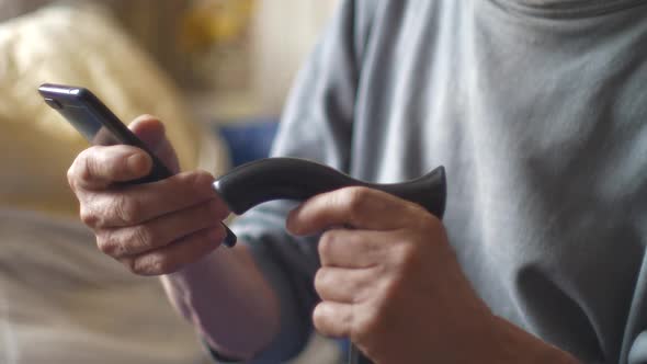 An elderly man with a walking stick uses a smartphone while sitting. The pensioner is holding a phon alt