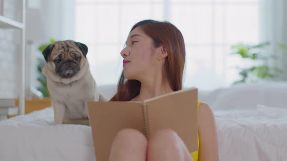 Beautiful Asian young woman reading book with dog in cozy bedroom alt
