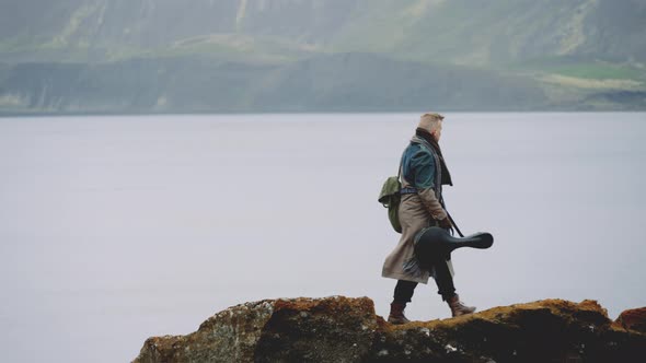 Man With Guitar Case And Greatcoat Walking Along Rocks By Valley River alt