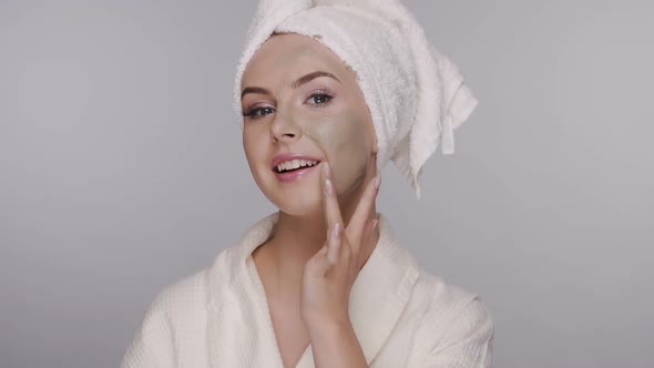 Girl in Bathrobe Applying Facial Clay Mask and Smoothes on Her Face alt