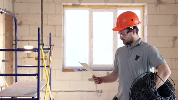 A builder in a helmet at a construction site examines a drawing and a work plan.