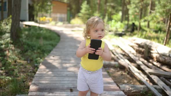 Little Funny Cute Blonde Girl Child Toddler Holding Playing Big Black Smartphone Outside at Summer