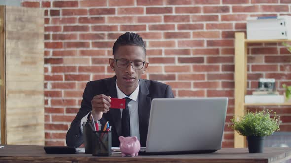 Black Man in Glasses and a Suit Enters the Bank Card Data Into a Laptop at the Office Table in alt