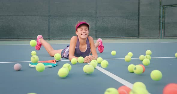 Cute Girl Laying on Tennis Court As a Bunch of Tennis Balls Rolling Towards Her alt