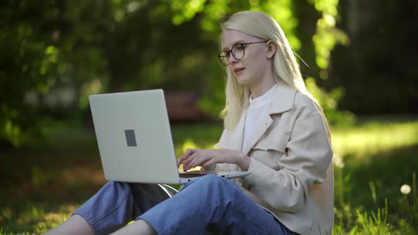 Young Blonde Woman in Glasses with Laptop on Green Grass