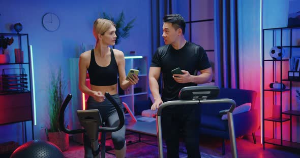 Woman and Asian Man Exercising at Home Together Using Running Track and Stationary Bike and Talking alt