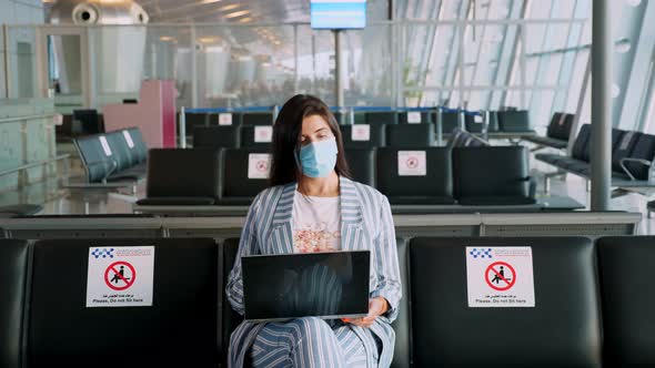 Woman, in Protective Mask, Uses Laptop, While Sitting in Empty Airport Lounge, Waiting To Board  alt