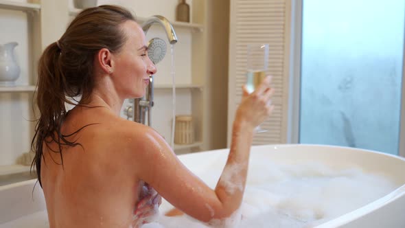 A Relaxed Woman Drinks Champagne From a Glass While Enjoying a Bubble Bath in the Bathtub alt