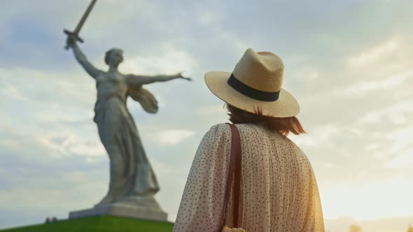A young girl in a dress looking at Mother Motherland on the Mamayev Kurgan at sunset alt